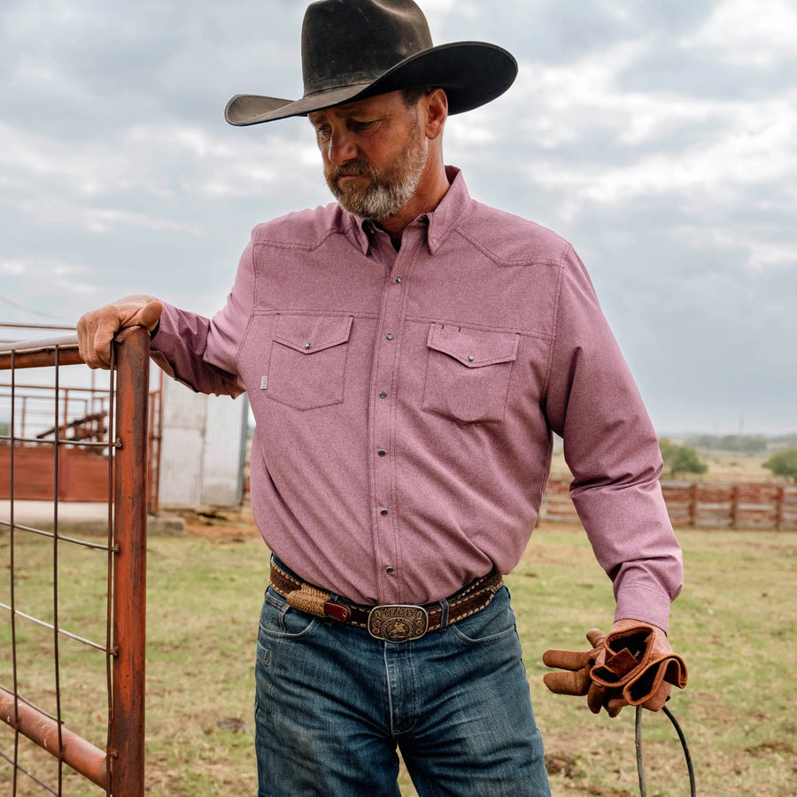 Man wearing a maroon pearl snap shirt, blue jeans and a black cowboy hat beside a ranch gate